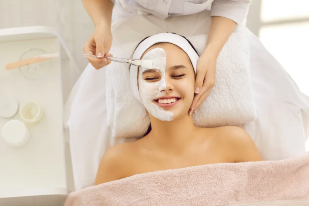 Smiling woman receiving a relaxing facial treatment at a medspa, with a brush applying a white mask.